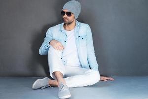 Relaxed and carefree. Handsome young man in eyewear sitting on the floor against grey background photo