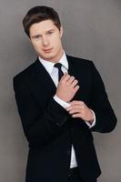 Young and successful. Handsome young man in formalwear adjusting his sleeve and looking at camera while standing against grey background photo