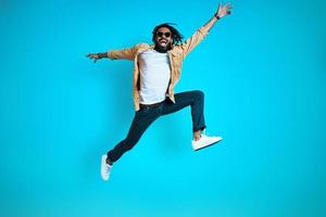 toda la longitud de un joven africano alegre con ropa informal gritando y gesticulando mientras se cierne sobre un fondo azul foto