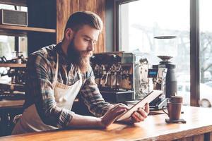 Technologies make business easier. Young bearded man in apron using his digital tablet while leaning to bar counter at cafe photo
