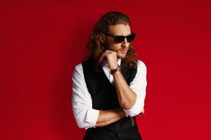 Confident young man in sunglasses looking away and keeping hand on chin while standing against red background photo