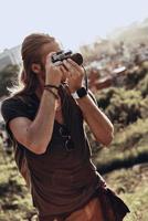 His passion. Young man in casual clothing photographing the view while standing on the hill outdoors photo