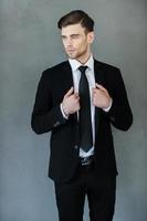 Looking perfect and trendy. Handsome young man adjusting his jacket and looking away while standing against grey background photo