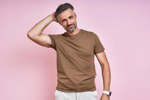 Handsome mature man holding hand behind head while standing against pink background photo