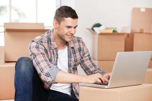 Searching a good moving company. Cheerful young man sitting on the floor and typing something on laptop while cardboard boxes laying on the background photo