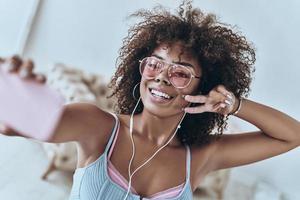 Having fun. Attractive young African woman listening music and smiling while taking selfie at home photo