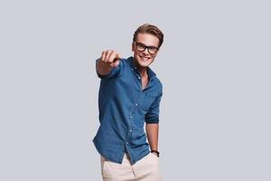 Handsome young man pointing at you and smiling while standing against grey background photo