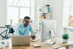 un apuesto hombre de mediana edad con ropa informal inteligente que usa una tableta digital y sonríe mientras está sentado en la oficina foto