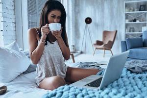 Morning starts with coffee and Internet. Attractive young smiling woman drinking coffee and looking at laptop while sitting on the bed at home photo