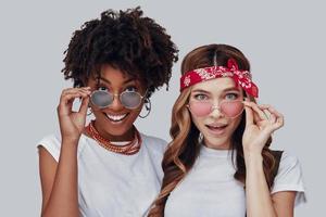 Two surprised young women looking at camera and smiling while standing against grey background photo