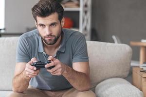 totalmente concentrado en su juego. apuesto joven jugando videojuegos mientras está sentado en el sofá en casa foto