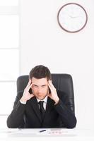 Time to take a break. Depressed young man in formalwear looking at camera and holding head in hands while sitting at his working place with wall clock on background photo