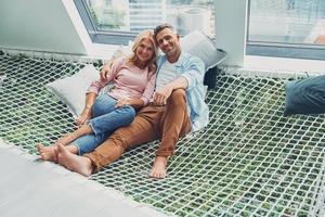 hermosa pareja madura sonriendo y mirando a la cámara mientras se relaja en una gran hamaca en casa juntos foto