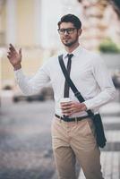 Hey taxi Confident young man in glasses holding coffee cup and waving for taxi while walking outdoors photo