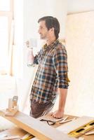 Sip of imagination. Side view of thoughtful young male carpenter holding coffee cup while leaning at the wooden table with diverse working tools laying on it photo