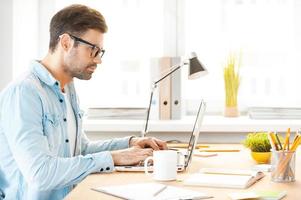 Youthful focus on great solution. Side view of handsome young man in shirt and eyewear working on laptop while sitting at his working place photo