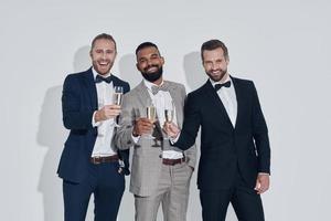 Three handsome young men in suits and bowties toasting with champagne while standing against gray background photo