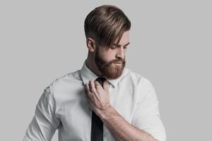 Thoughtful businessman. Young beautiful man adjusting his necktie and looking away while standing against grey background photo