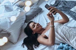 Chatting with boyfriend. Top view of attractive young woman using smart phone and smiling while lying on the bed at home photo