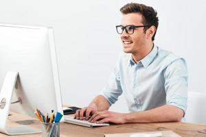 gran solución todos los días. joven apuesto alegre con anteojos trabajando en la computadora y sonriendo mientras se sienta en su lugar de trabajo foto