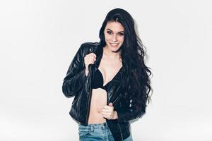 Beautiful seductress. Beautiful young woman taking off her leather jacket and smiling while standing against grey background and smiling photo