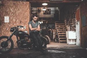 hombre cerca del garaje de motocicletas. un joven guapo con los brazos cruzados y mirando pensativo mientras se apoya en su bicicleta con un taller de reparación en el fondo foto