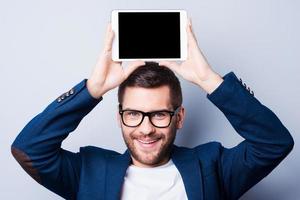 Copy space on his tablet. Cheerful young man holding a digital tablet upon his head and smiling while standing against grey background photo