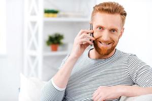 Lovely communication. Happy bearded man talking on the mobile phone and smiling at camera while sitting on sofa photo