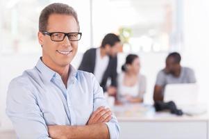 Confident in his team. Confident mature man in glasses keeping arms crossed and smiling while three people working on background photo