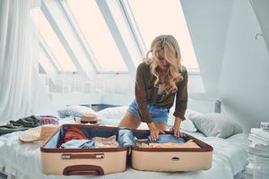 bella joven empacando equipaje mientras se sienta en la cama en casa foto