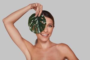 Happiness highlights her beauty. Attractive young woman looking at camera and covering face with leaf while standing against grey background photo
