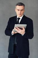Technologies make business successful. Mature businessman using his touchpad while standing against grey background photo