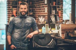 dueño de la barbería. alegre joven barbudo mirando a la cámara y sosteniendo la mano en el bolsillo mientras se apoya en una silla en la barbería foto