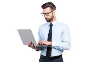 Concentrated on work. Confident young handsome man in shirt and tie working on laptop while standing against white background photo