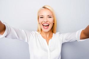 Happy moments must be saved. Cheerful young blond hair woman holding camera and making photo of herself while standing against grey background