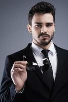 Stylish handsome. Portrait of confident young man in formalwear holding sunglasses and looking at camera while standing against grey background photo