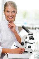Smart and confident. Smiling young female scientist writing in note pad and looking at camera while sitting at her working place photo