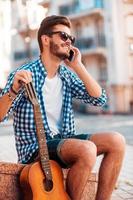 esperando amigos. vista de ángulo bajo de un joven alegre sosteniendo guitarra y hablando por teléfono móvil mientras se sienta al aire libre foto