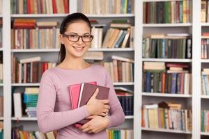 sintiéndose segura de sus exámenes. alegre joven estudiante sosteniendo libros y sonriendo mientras está de pie en la biblioteca foto