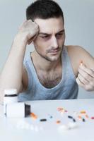 Numbing his pain. Frustrated young man sitting at the table and looking at the pills in his hand while more pills laying on the table photo