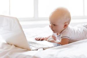 Baby from digital age. Cute little baby holding hands on laptop keyboard and looking at monitor while lying in bed photo