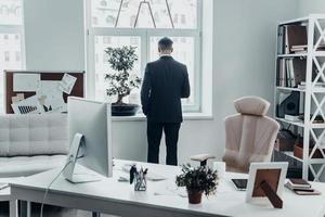 Hard working day. Rear view of young man in full suit standing near the window in the office photo