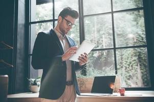 Working on new tablet. Low angle view of confident young man working on digital tablet while standing in front of the big window in office or cafe photo