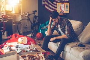 Terrible hangover. Frustrated young man keeping hand in hair while sitting on sofa in messy room after party photo