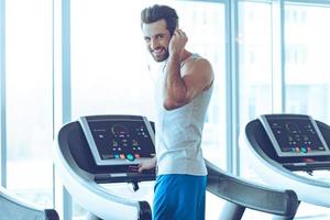 What did you say Rear view of young handsome man in sportswear standing on treadmill in front of window at gym and adjusting his headphone while looking at camera with smile photo
