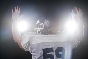 Welcoming stadium.  Rear view of American football player holding football ball keeping arms raised while standing against lights photo