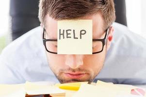 He needs help. Frustrated young man in formalwear with adhesive note on his forehead leaning his head at the table photo