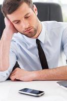 Waiting for call. Depressed young man in shirt and tie holding head in hand and looking at the mobile phone laying on the table photo