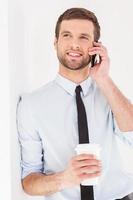 Having a coffee break. Cheerful young man in shirt and tie talking on the mobile phone and drinking coffee while leaning at the wall photo