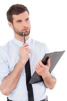 Businessman with clipboard. Thoughtful young man in formalwear holding clipboard and touching his chin with pen while standing isolated on white background photo